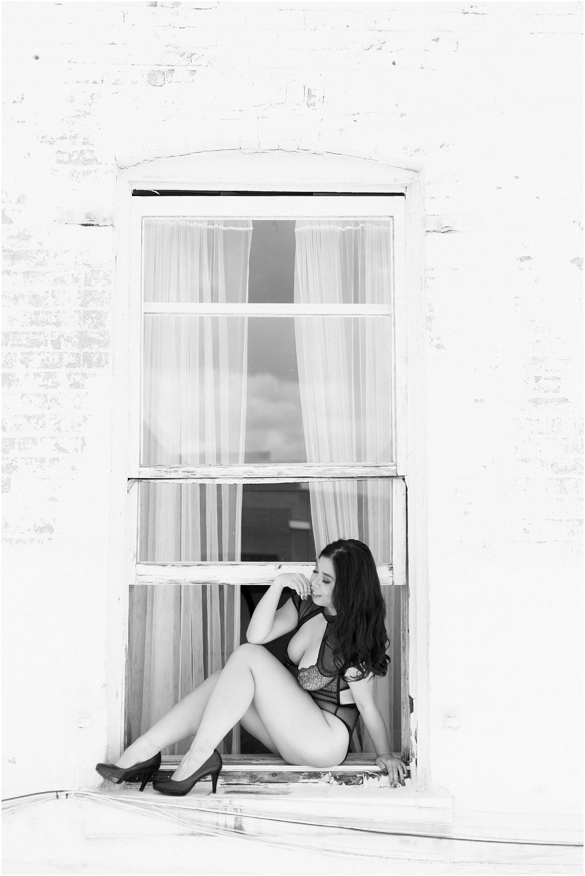 Far away black and white photo of a model in black lingerie sitting on a window sill. 
