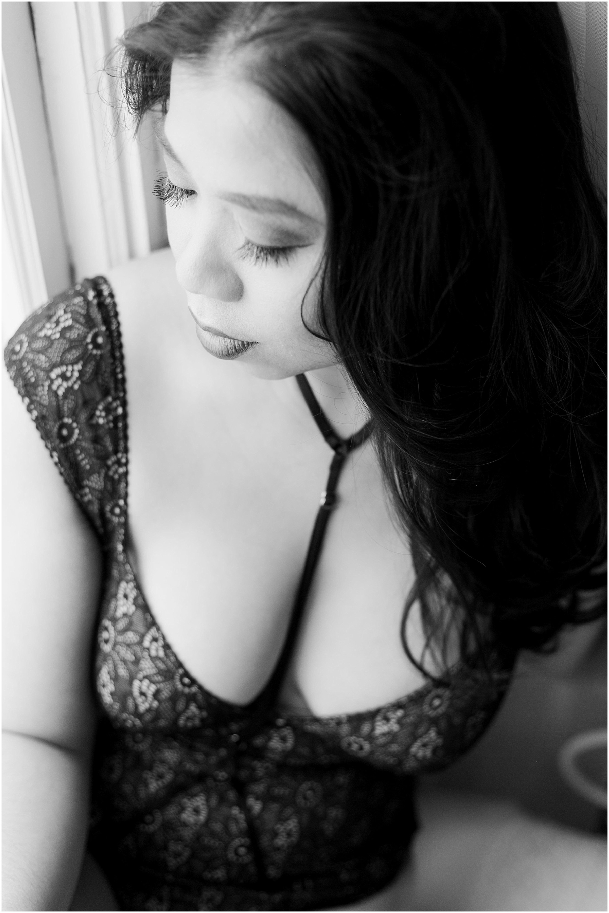Black and white photo of a lady standing next to the window wearing black lace lingerie. 