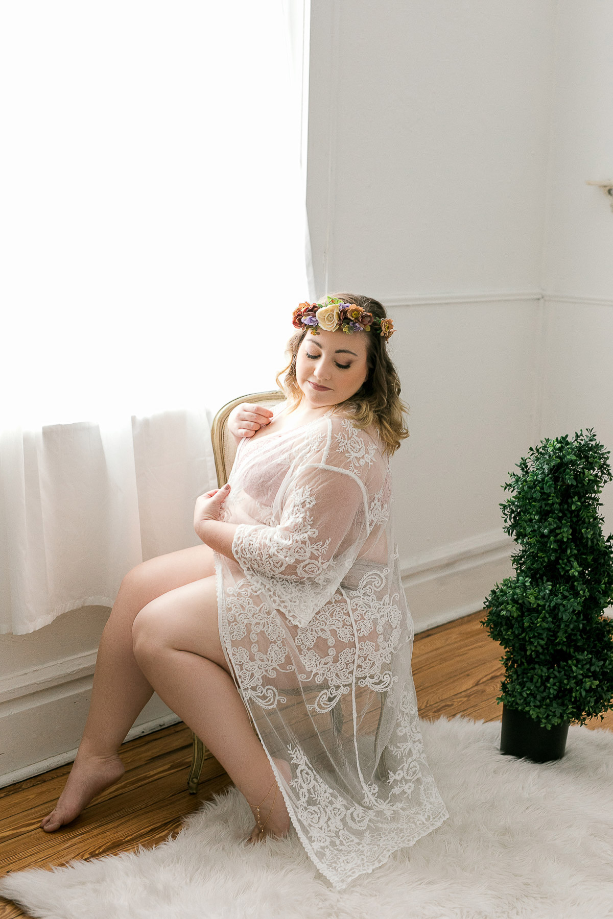 Curvy woman in boudoir shoot sitting in front of the window looking at her lace robe.