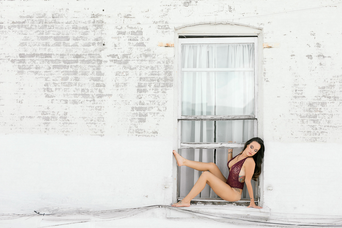 Woman sitting at a window outside in maroon lingerie bodysuit. 