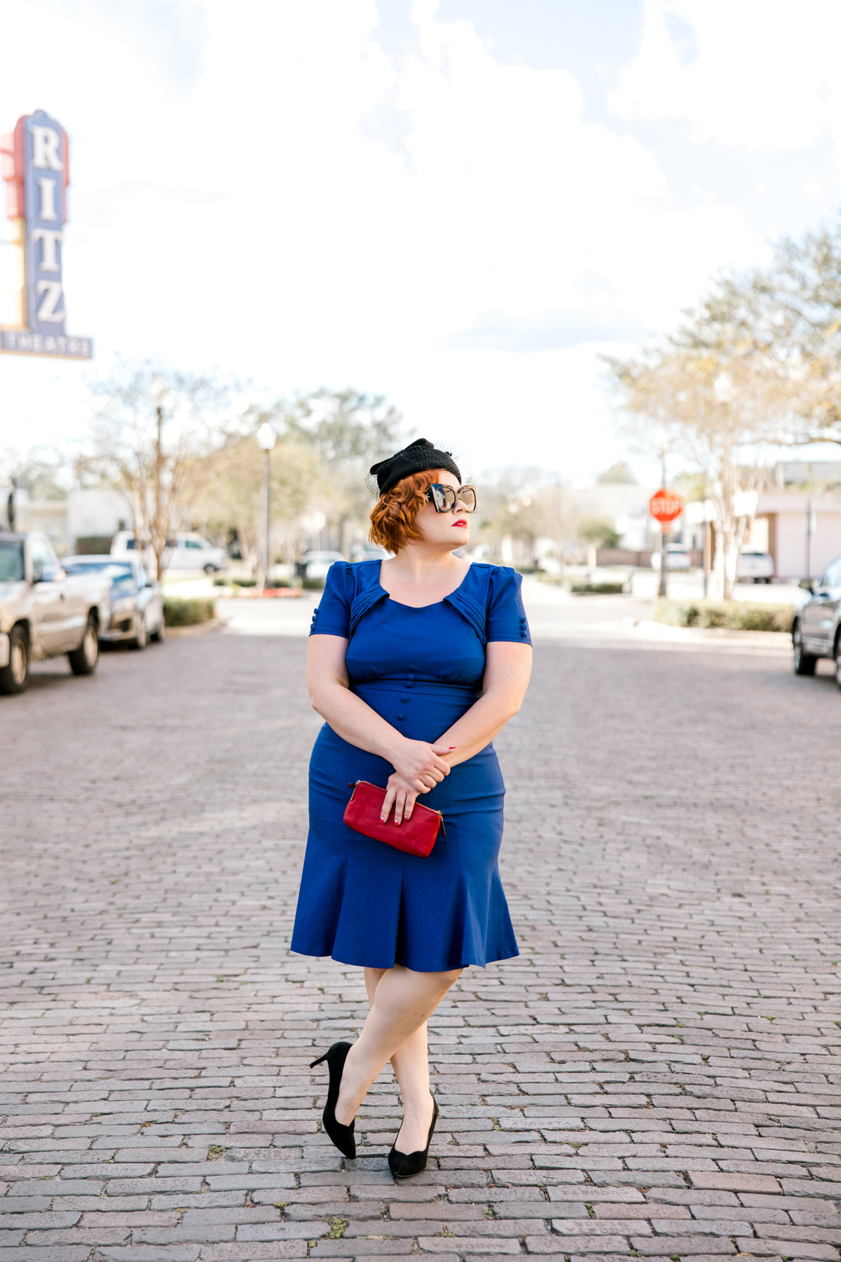 Old Hollywood glam 1950's photo shoot -- woman in vintage blue dress, pillbox hat, red purse, black pumps -- big sunglasses