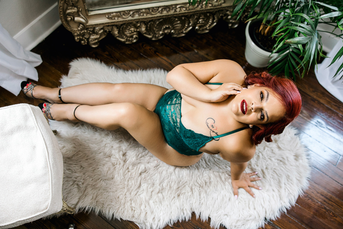 stunning red head in green lacy bodysuit and heels on white fur rug looking up at the camera