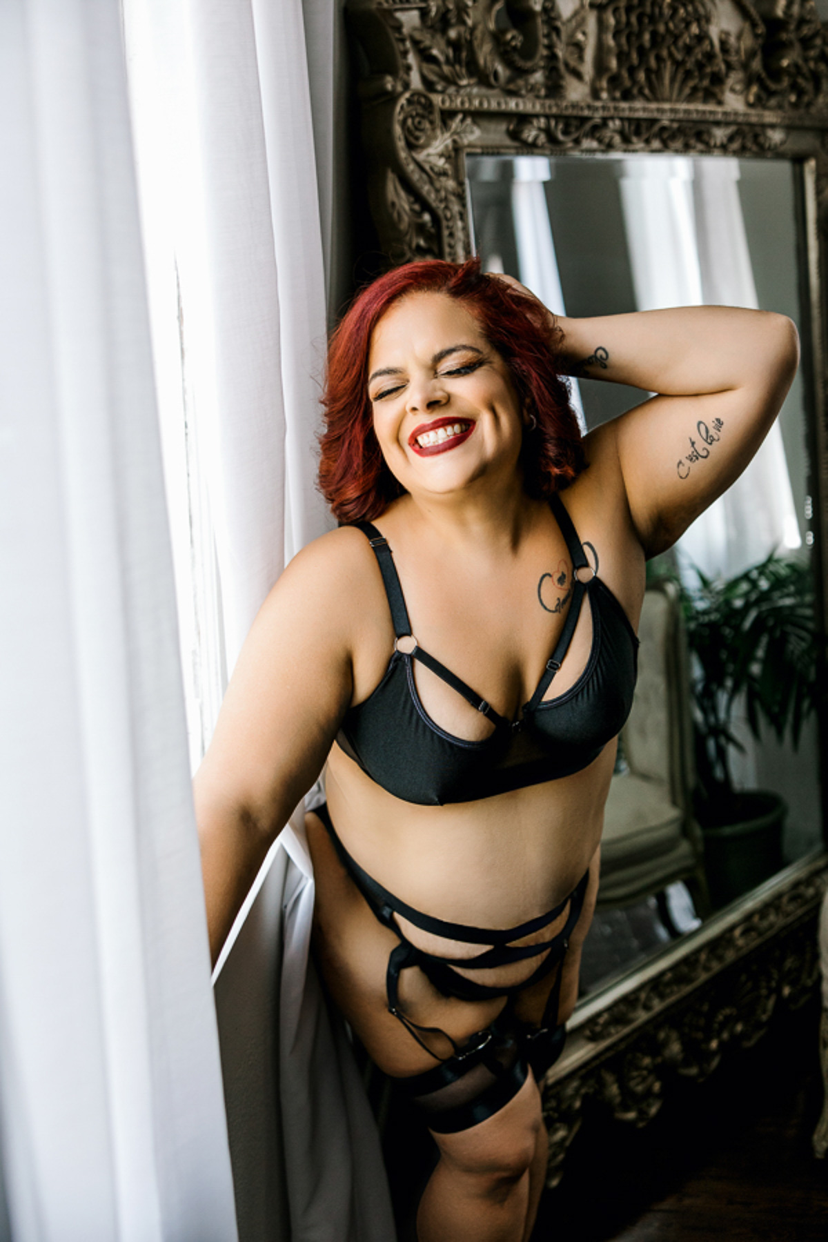 curvy girl with red hair in black bra and panty and garter set smiling in front of window sexy & empowered